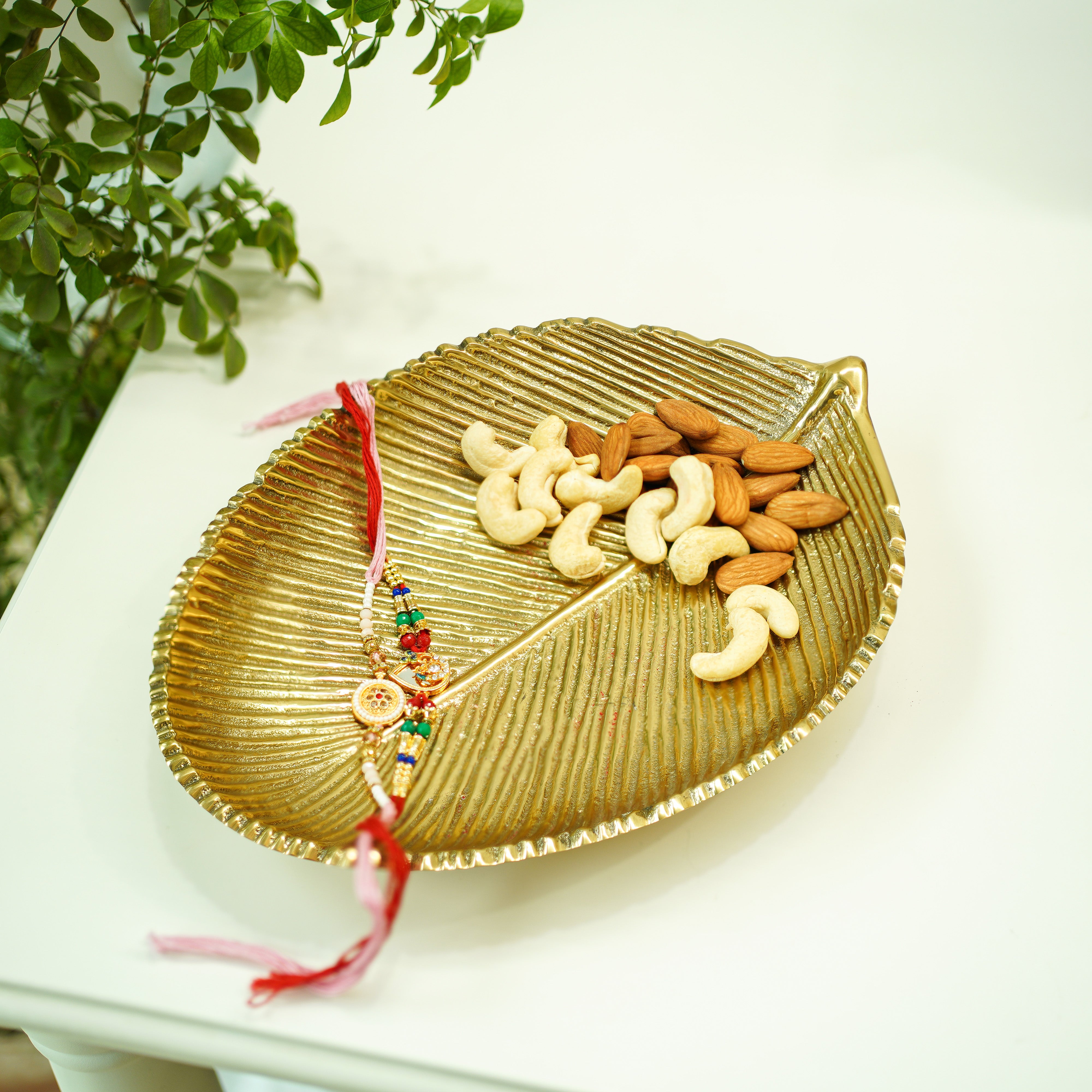Leaf-shaped gold tray with nuts and a colorful Rakhi on a white surface with greenery.