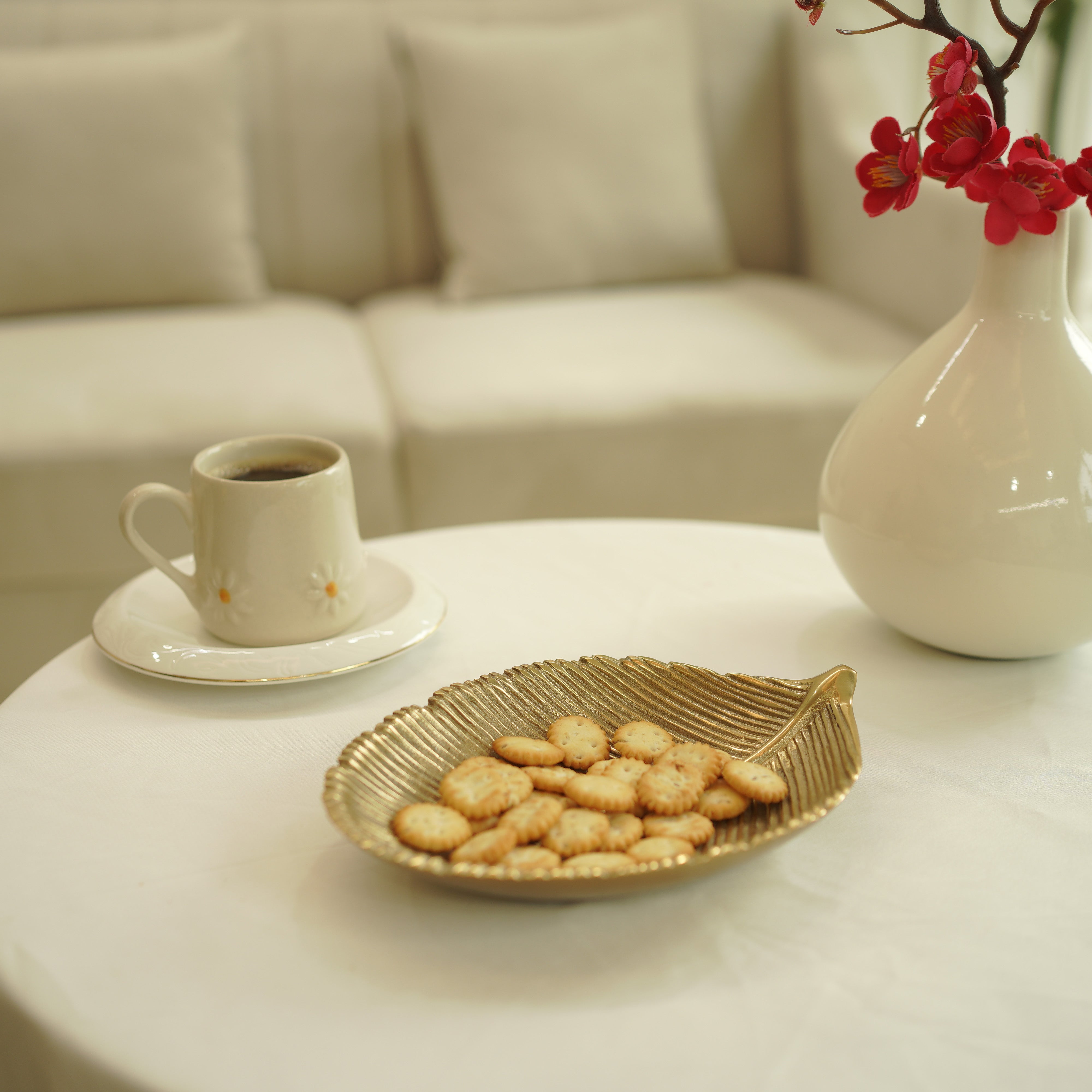 Cup of coffee with cookies on a leaf-shaped plate, vase with red flowers in the background