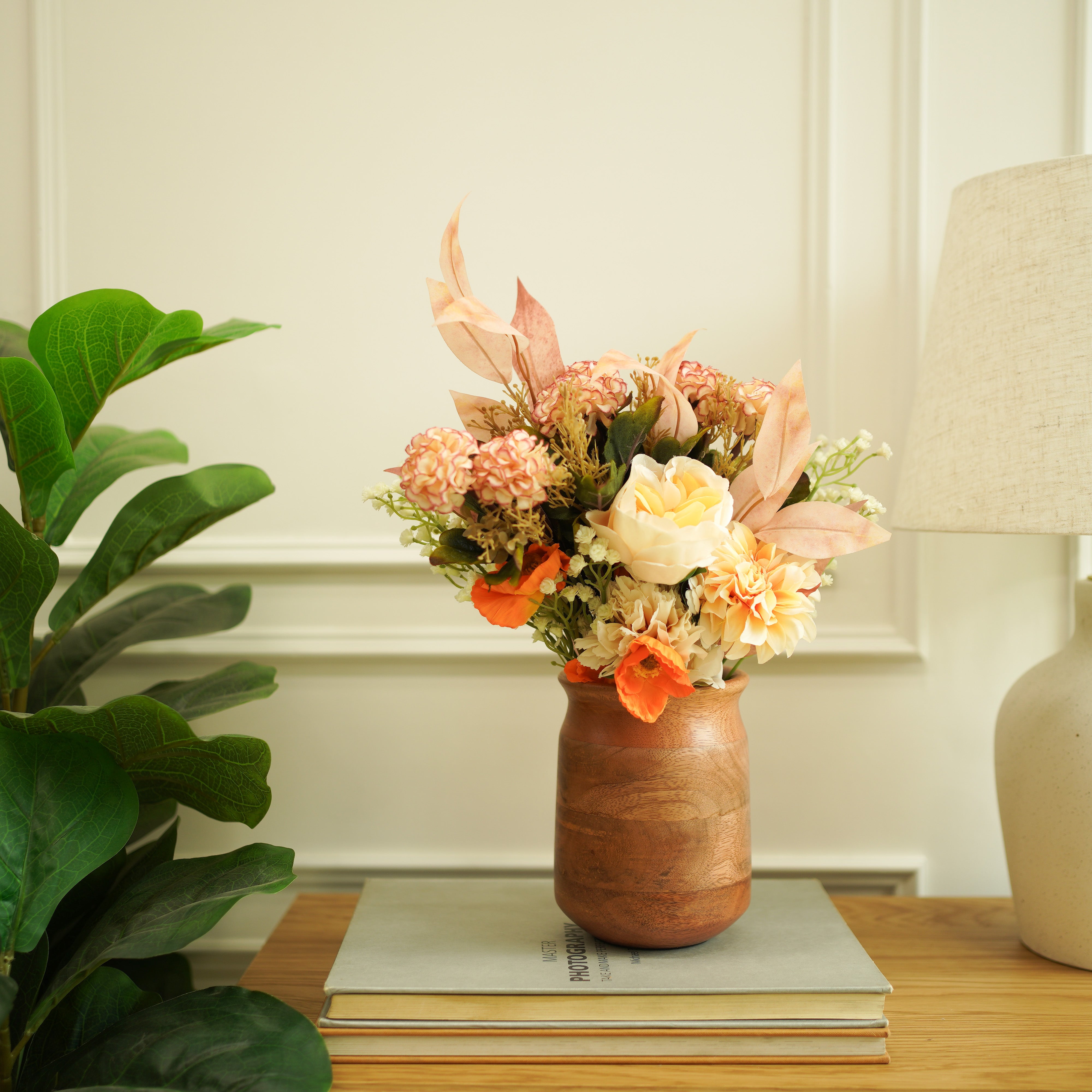 Wooden vase with flowers on a stack of books next to a plant and lamp.