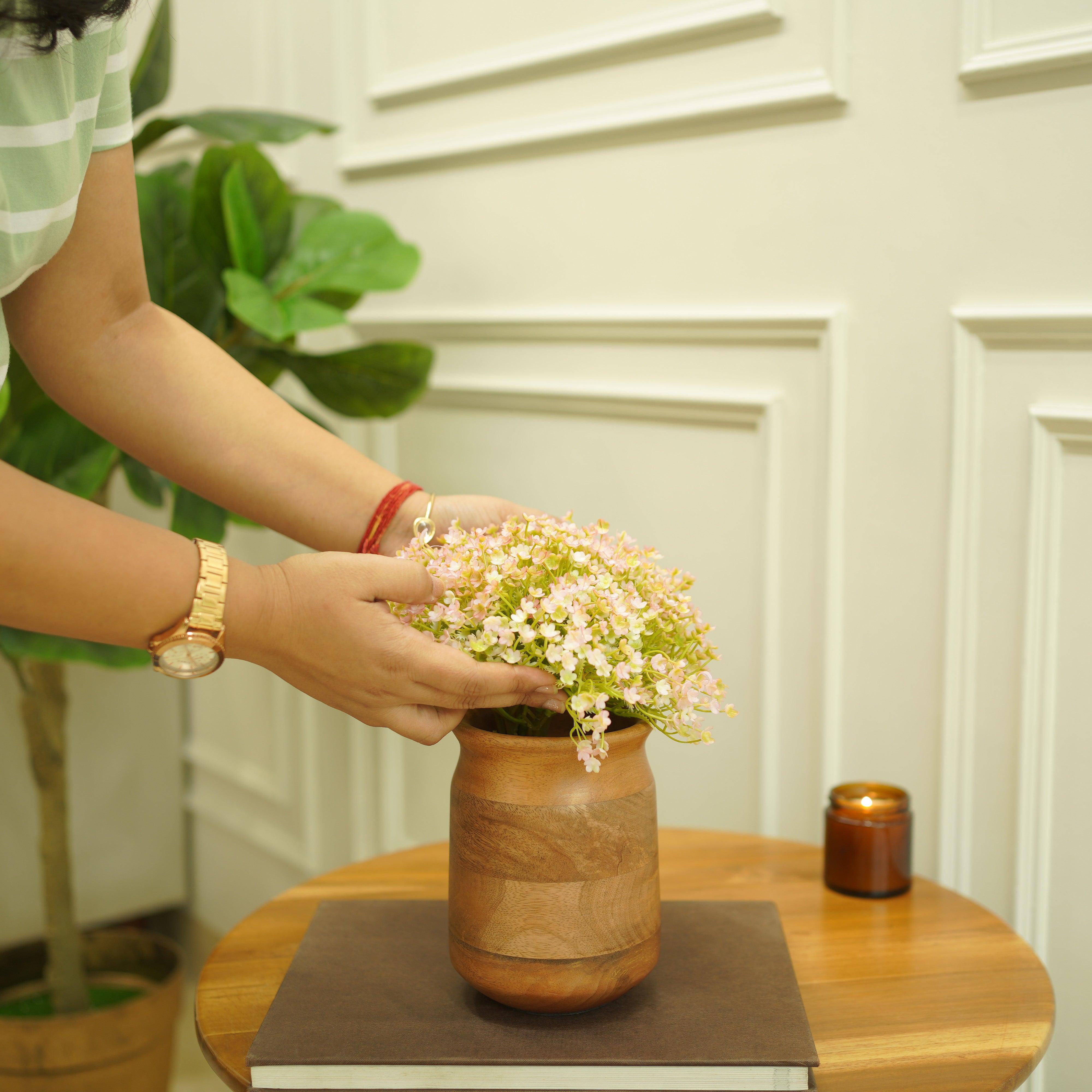 Person arranging flowers in a wooden mango vase on a table with a white paneled wall in the background.