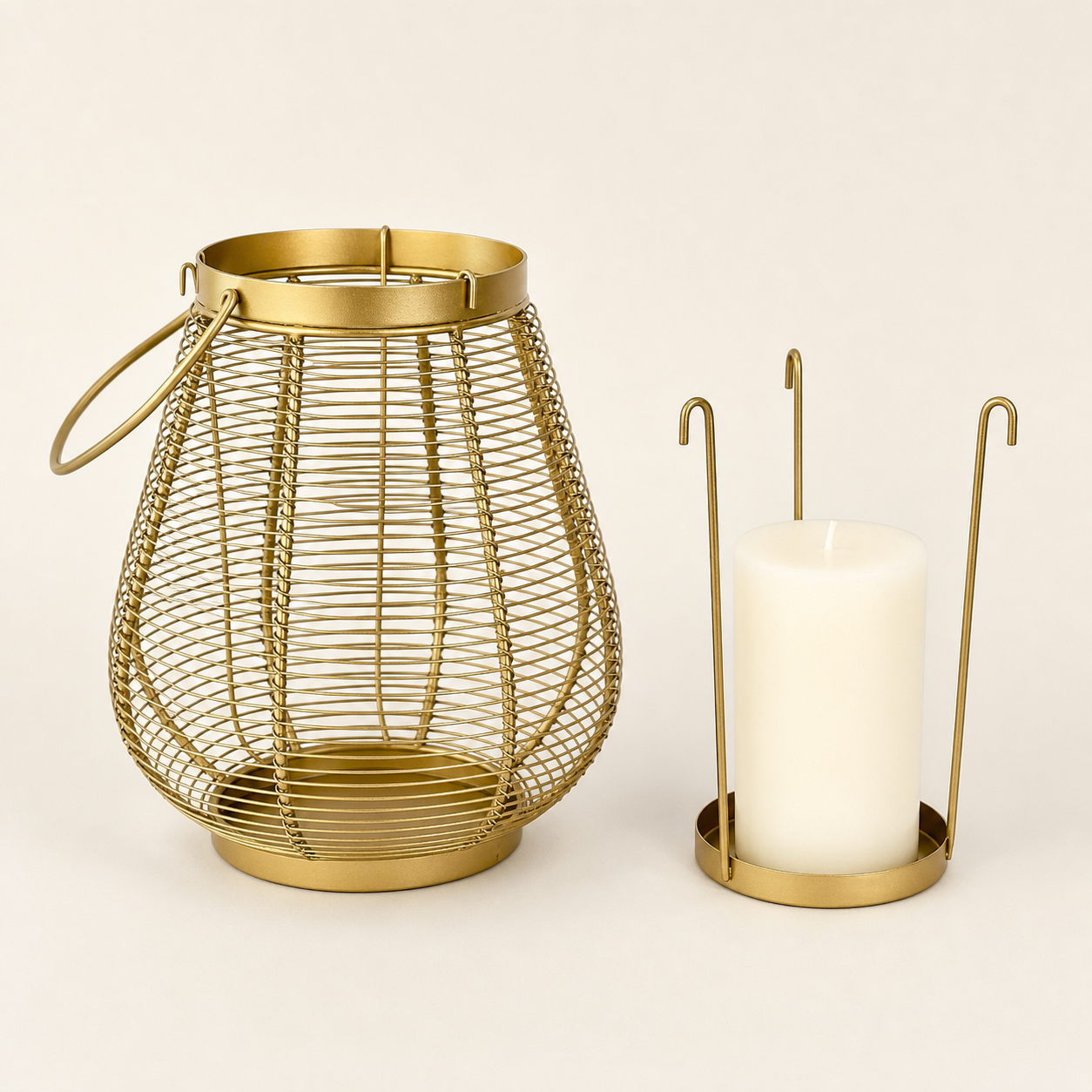 A gold-toned metal wire lantern with a top handle sits next to a white pillar candle in a matching gold holder, set against a plain light beige background.