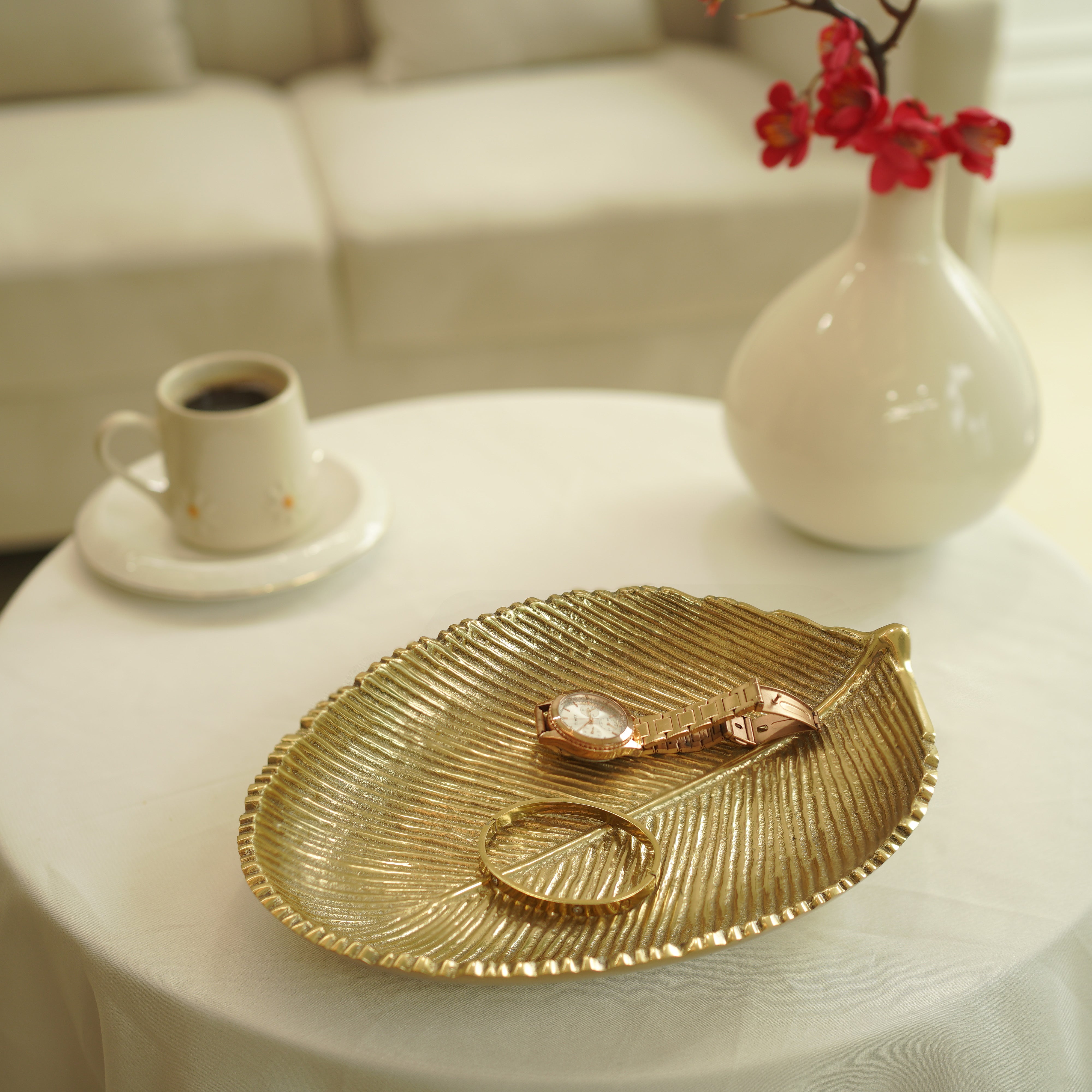 Gold leaf-shaped tray with jewelry on a white table, coffee cup, and vase in the background.
