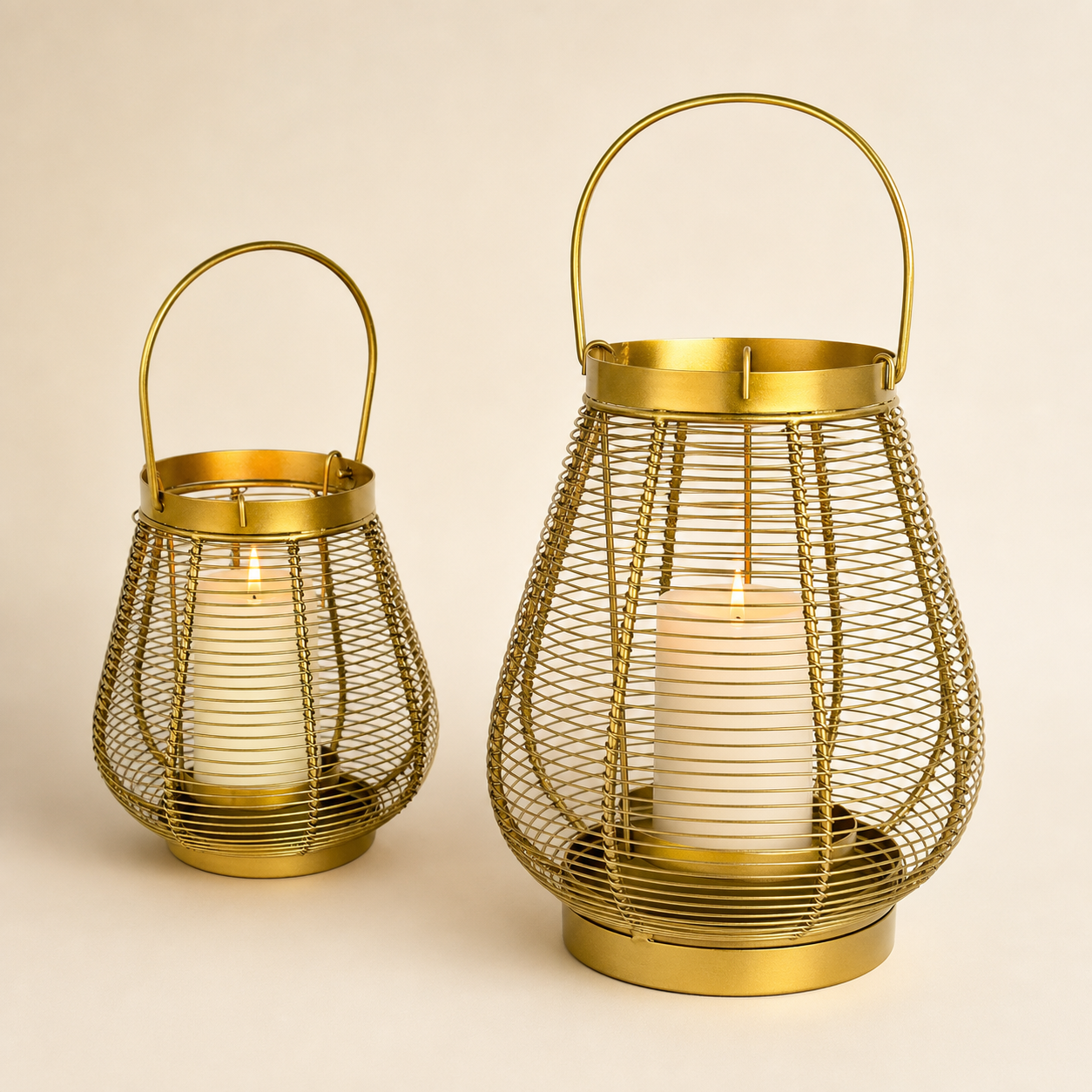 Two gold metal wire lanterns with arched handles, each containing a white pillar candle, photographed on a plain warm beige background.